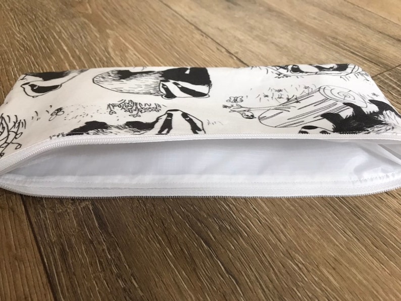 May include: A white zippered pouch with a black and white print of badgers. The pouch is lying flat on a wooden surface.
