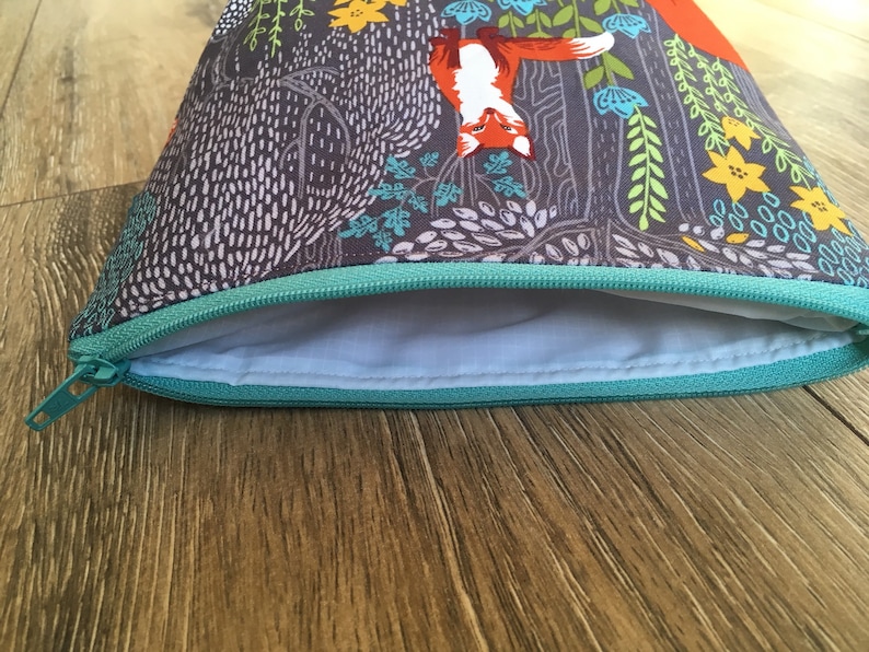 May include: A gray zippered pouch with a teal zipper and a colorful fox print. The print features orange foxes, green leaves, and yellow flowers.
