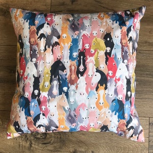 May include: A square cushion featuring a repeating pattern of horse faces in a variety of colours. The design includes shades of white, black, brown, and blue. The cushion appears to be made of fabric.