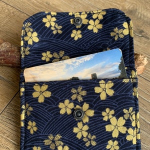 May include: A navy blue card holder with a gold floral pattern and a snap closure. The interior holds a card with a landscape image. The holder is open, revealing the card inside. The background is a wooden surface.