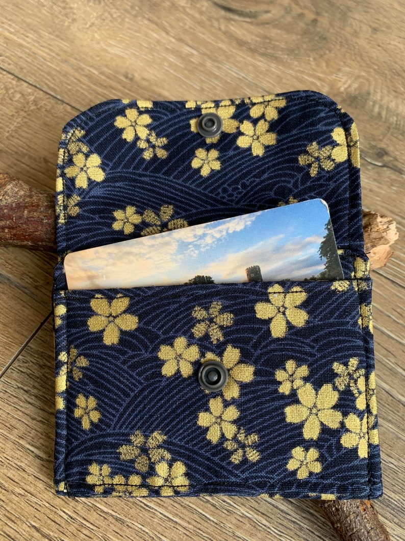 May include: A navy blue card holder with a gold floral pattern and a snap closure. The interior holds a card with a landscape image. The holder is open, revealing the card inside. The background is a wooden surface.