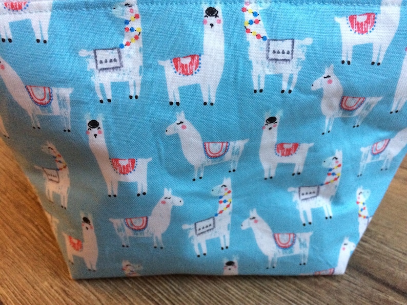 May include: A blue fabric tote bag with a repeating pattern of white llamas with colorful accents. The llamas are wearing saddles and have different colored headbands.