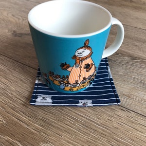 K&ouml;nnte beinhalten: Eine blaue Keramiktasse mit wei&szlig;em Innenraum und einer Cartoon-Illustration eines Charakters mit einer Gruppe kleinerer Charaktere. Die Tasse steht auf einem blauen und wei&szlig;en gestreiften Stoffuntersetzer.