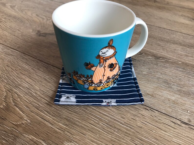 K&ouml;nnte beinhalten: Eine blaue Keramiktasse mit wei&szlig;em Innenraum und einer Cartoon-Illustration eines Charakters mit einer Gruppe kleinerer Charaktere. Die Tasse steht auf einem blauen und wei&szlig;en gestreiften Stoffuntersetzer.