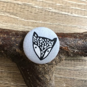 May include: A circular, pale blue fabric button featuring a black and white fox face design. The fox has a white face and black outlines. The button is on a brown branch.