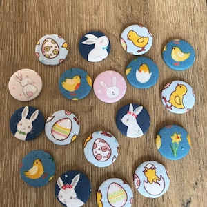 May include: A collection of 17 Easter-themed fabric buttons featuring bunnies, chicks, and eggs. The buttons are in shades of blue, pink, yellow, and white.