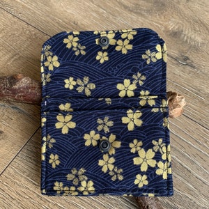 May include: Open navy blue card holder with a gold floral pattern. The fabric wallet features a snap closure and is displayed open, revealing the interior. The design includes a wave pattern.