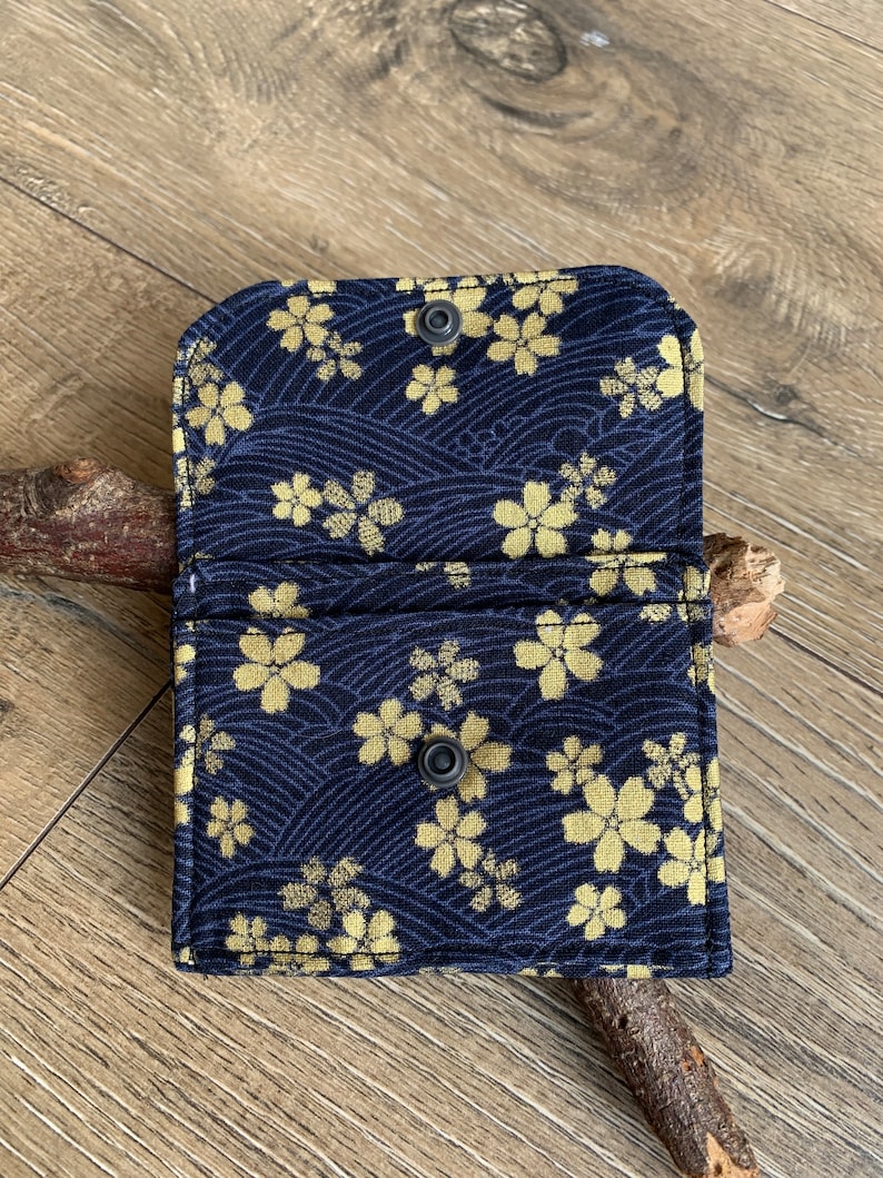 May include: Open navy blue card holder with a gold floral pattern. The fabric wallet features a snap closure and is displayed open, revealing the interior. The design includes a wave pattern.