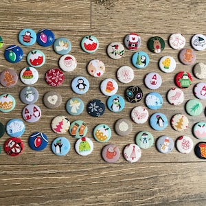May include: A collection of 48 round Christmas-themed buttons with various designs, including penguins, snowmen, reindeer, and Christmas trees. The buttons are all different colors and have a variety of textures.