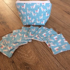 Op de afbeelding: Een lichtblauwe make-uptas en bijpassende herbruikbare wattenschijfjes. De tas en de schijfjes hebben een herhalend patroon van witte lama's met rode en roze accenten. De tas heeft een witte ritssluiting.