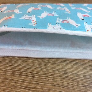 May include: A light blue zippered pouch with a white zipper and a white lining. The pouch is decorated with a repeating pattern of white llamas with rainbow accents.