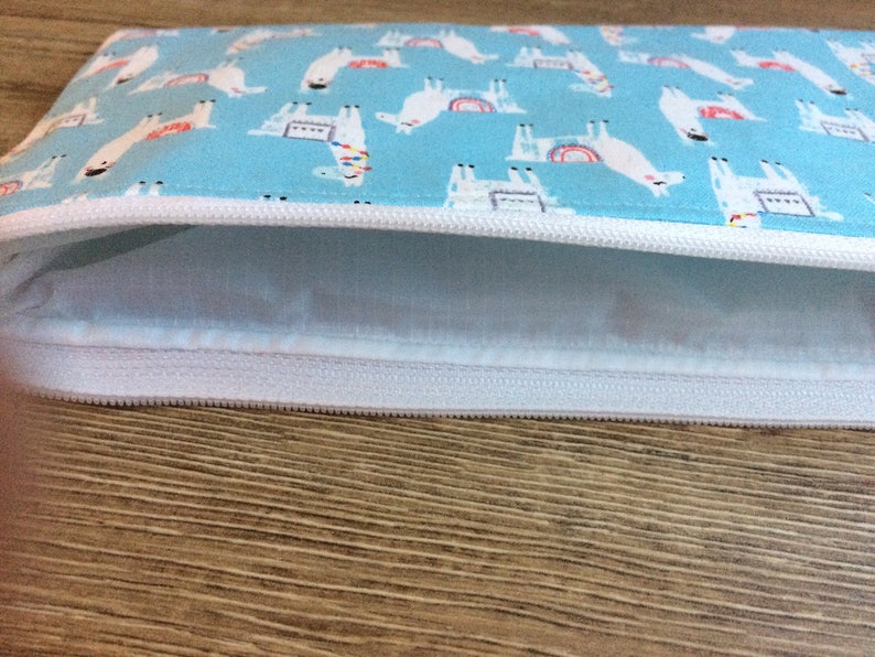 May include: A light blue zippered pouch with a white zipper and a white lining. The pouch is decorated with a repeating pattern of white llamas with rainbow accents.