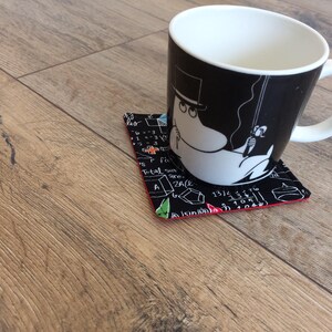 May include: A black and white mug with a cartoon character on it, sitting on a black and white coaster with red trim. The coaster has a pattern of mathematical equations and symbols.