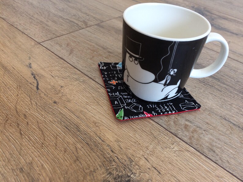 May include: A black and white mug with a cartoon character on it, sitting on a black and white coaster with red trim. The coaster has a pattern of mathematical equations and symbols.