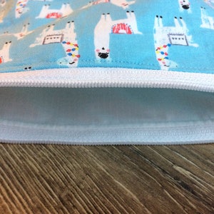 May include: A light blue zippered pouch with a white zipper and a white lining. The pouch is decorated with a repeating pattern of white llamas with colorful accents on a light blue background.