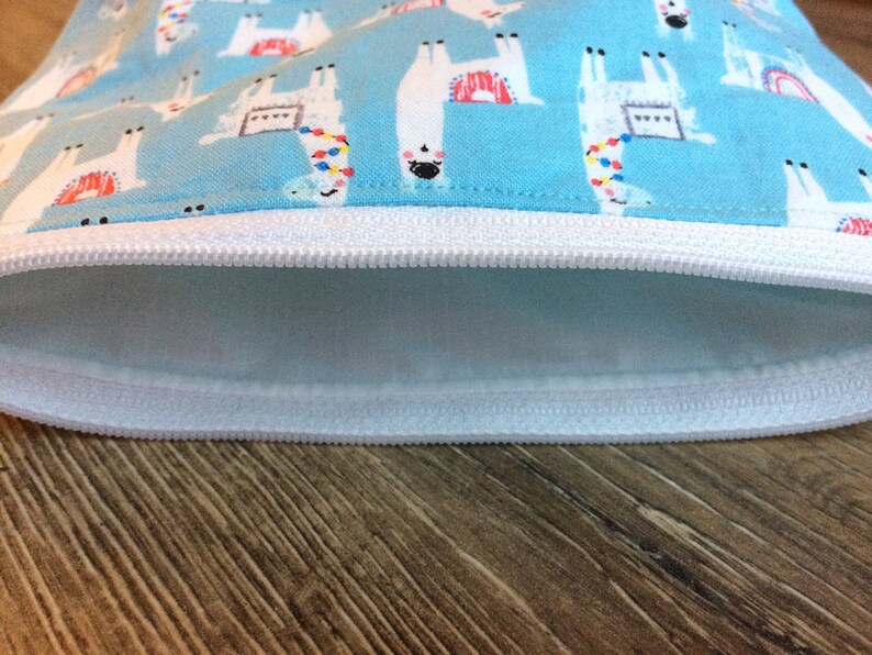May include: A light blue zippered pouch with a white zipper and a white lining. The pouch is decorated with a repeating pattern of white llamas with colorful accents on a light blue background.