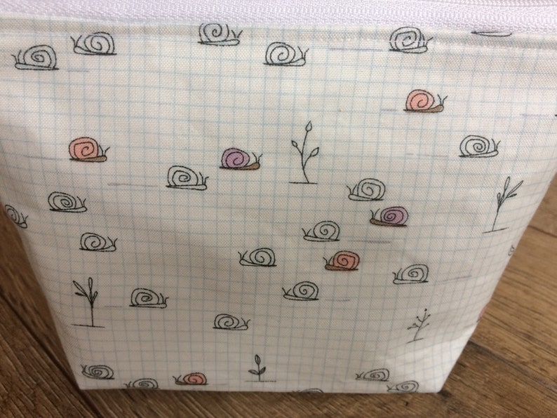 Pu&ograve; includere: Una pochette bianca con cerniera, con un motivo a griglia e un design ripetitivo di lumache nere. Alcune delle lumache hanno conchiglie rosa o viola.