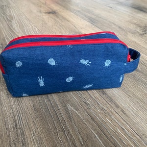 Può includere: Un astuccio per matite in denim blu con una cerniera rossa e un motivo di scarabei bianchi. L'astuccio ha un manico.