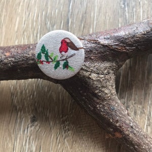 Peut inclure: Un bouton rond en tissu avec un rouge-gorge perché sur une branche avec des baies rouges. Le bouton est beige avec un motif vert et rouge.