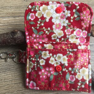 以下が含まれることがあります： 花柄の赤い布製の財布で、スナップ留めが2つ付いています。財布には銀色のキーチェーンが付いています。