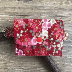 May include: A small, red fabric wallet with a floral pattern of pink and white blossoms and gold accents. The wallet has a snap closure and a silver clasp attached to a matching strap. The background is a wooden surface.