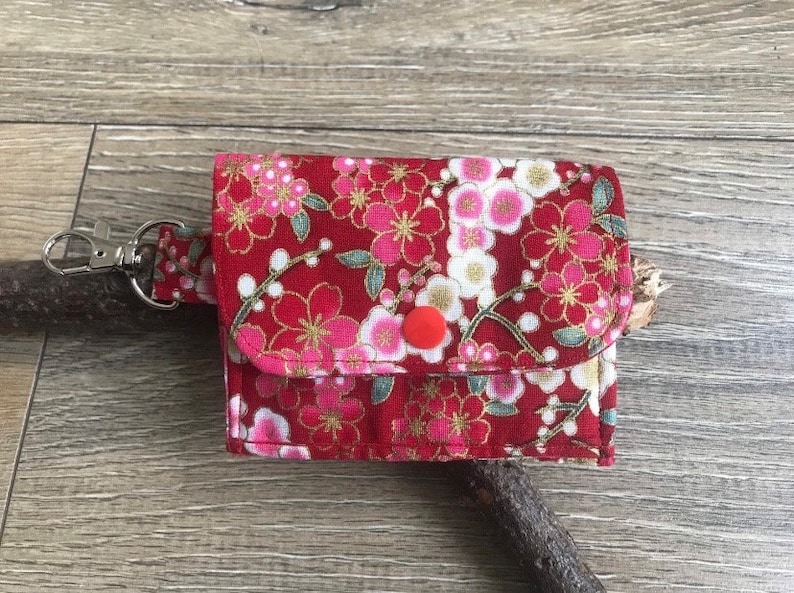 May include: A small, red fabric wallet with a floral pattern of pink and white blossoms and gold accents. The wallet has a snap closure and a silver clasp attached to a matching strap. The background is a wooden surface.