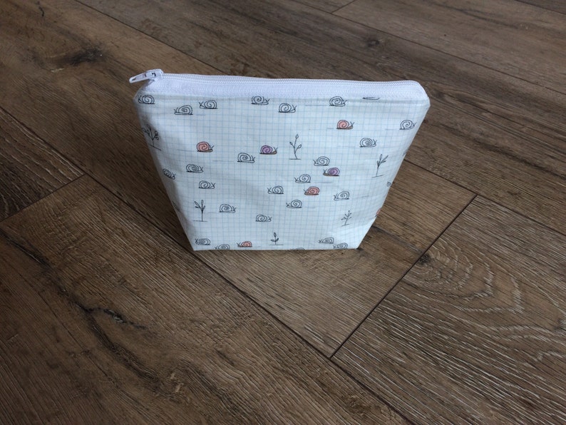 Pu&ograve; includere: Borsa bianca con cerniera, motivo a griglia e un motivo ripetitivo di piccole lumache nere.