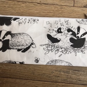 Könnte beinhalten: Eine weiße Reißverschlusstasche mit einem schwarz-weißen Dachs-Druck. Der Dachs ist in verschiedenen Posen dargestellt, darunter beim Gehen, Spielen und Kuscheln. Die Tasche ist perfekt zum Verstauen kleiner Gegenstände wie Stifte, Bleistifte und Make-up.