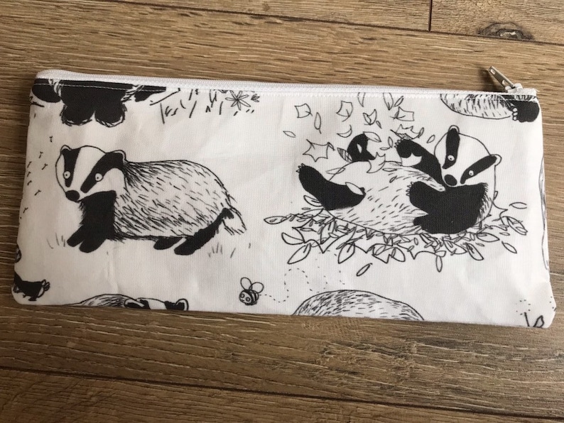 May include: A white zippered pouch with a black and white badger print. The badger is depicted in various poses, including walking, playing, and cuddling. The pouch is perfect for storing small items like pencils, pens, and makeup.