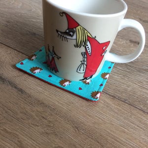 Pu&ograve; includere: Una tazza in ceramica bianca con un'illustrazione a cartoni animati di una donna con un vestito rosso e un cappello bianco. La tazza &egrave; appoggiata su un sottobicchiere blu turchese con un bordo rosso e un motivo di piccoli ricci marroni.