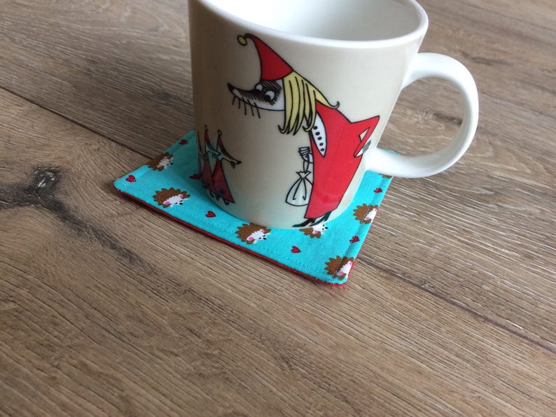 Pu&ograve; includere: Una tazza in ceramica bianca con un'illustrazione a cartoni animati di una donna con un vestito rosso e un cappello bianco. La tazza &egrave; appoggiata su un sottobicchiere blu turchese con un bordo rosso e un motivo di piccoli ricci marroni.