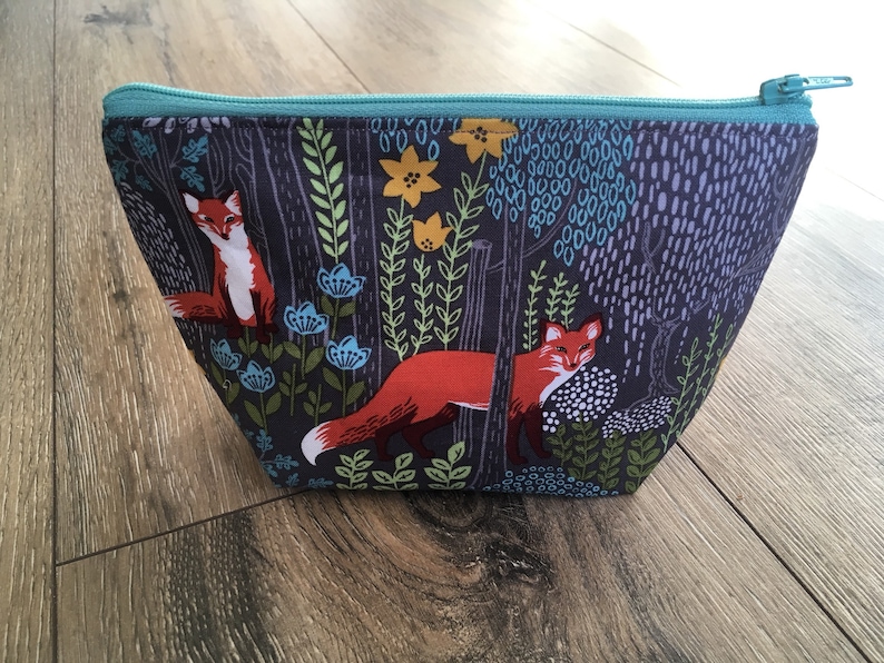 May include: A teal zippered pouch with a dark gray background and a repeating pattern of foxes, flowers, and trees. The foxes are orange and white.