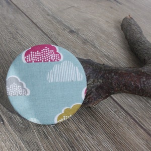 May include: A round, blue fabric pin with a white, grey, and pink cloud design. The pin has a yellow and white flower design in the bottom right corner.