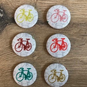 Peut inclure: Six boutons ronds en tissu avec un fond blanc et un motif de deux bicyclettes de différentes couleurs. Les bicyclettes sont dessinées en noir et les couleurs sont jaune, rouge, turquoise, or, rose et rouge.