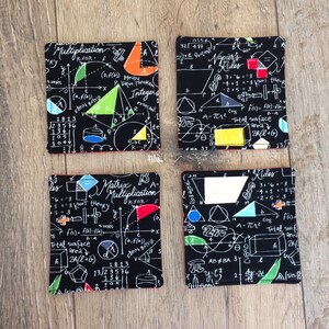 May include: Four black fabric coasters with a white mathematical formula and geometric shapes printed on them. The coasters have red stitching around the edges.