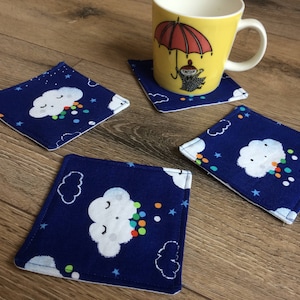 Puede incluir: Cuatro posavasos de tela azul con nubes blancas y gotas de lluvia de colores. Los posavasos tienen un borde blanco y están decorados con estrellas blancas.