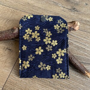 May include: A navy blue fabric case with a gold floral pattern and a black snap closure. The case features a repeating design of stylized flowers and wave-like lines. The case is likely for small items.