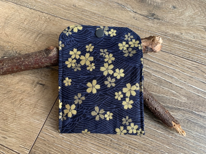 May include: A navy blue fabric case with a gold floral pattern and a black snap closure. The case features a repeating design of stylized flowers and wave-like lines. The case is likely for small items.