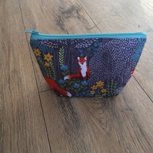 May include: A blue zippered pouch with a forest scene print featuring foxes, flowers, and trees. The pouch is made of fabric and has a teal zipper.