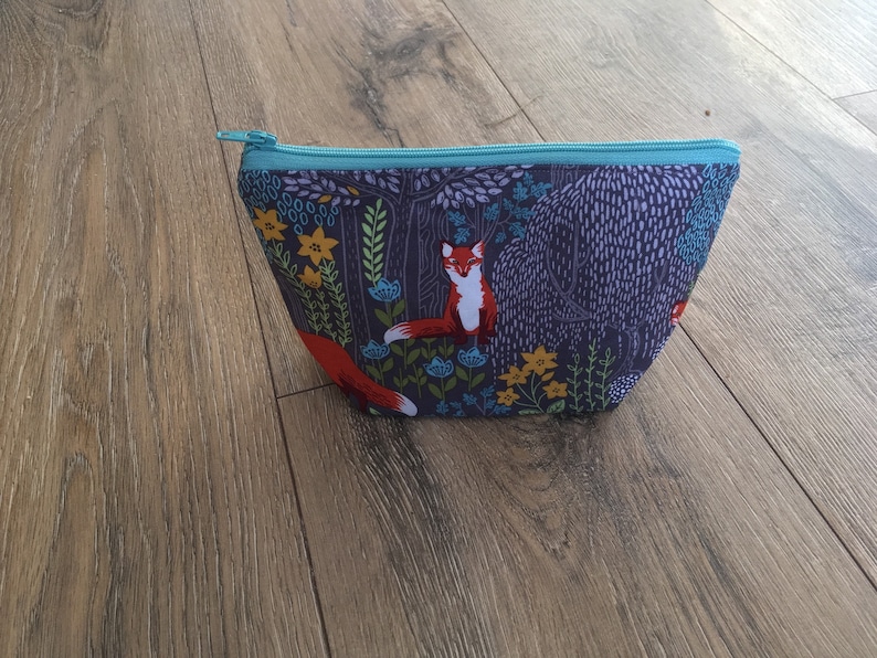 May include: A blue zippered pouch with a forest scene print featuring foxes, flowers, and trees. The pouch is made of fabric and has a teal zipper.