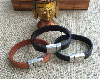May include: Three leather bracelets with silver magnetic clasps. The bracelets are brown, black, and black. The bracelets are arranged on a burlap surface in front of a golden statue of Buddha.