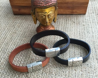 May include: Three leather bracelets with silver magnetic clasps. The bracelets are brown, black, and black. The bracelets are arranged on a brown textured surface.