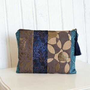 Könnte beinhalten: Eine braune und blaue Patchwork-Clutch mit einem schwarzen Quastenanhänger. Die Tasche hat ein geometrisches Muster und ein Blumenmuster.