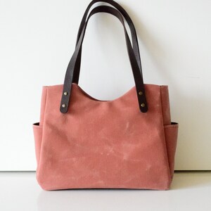 K&ouml;nnte beinhalten: Eine staubrosa Tragetasche mit dunkelbraunen Ledergriffen und Akzenten. Die Tasche hat zwei Au&szlig;entaschen und besteht aus einem robusten Canvas-Material. Die Griffe sind mit messingfarbenen Nieten befestigt.