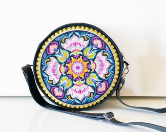May include: A round black crossbody bag with a colorful floral embroidery design. The bag has a long adjustable strap.