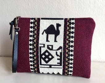 May include: A burgundy clutch purse with a white and black geometric pattern featuring a camel silhouette. The purse has a black leather strap.