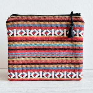May include: A rectangular, zippered pouch with a colorful woven pattern. The design features horizontal stripes in red, blue, yellow, and brown, with white geometric accents. A black tassel hangs from the zipper.