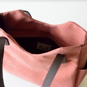 K&ouml;nnte beinhalten: Eine rosafarbene Tragetasche mit dunkelbraunen Ledergriffen und -besatz. Die offene Tasche zeigt ein dunkles Innenfutter und Seitentaschen. Die Tasche hat ein schlichtes, klassisches Design.