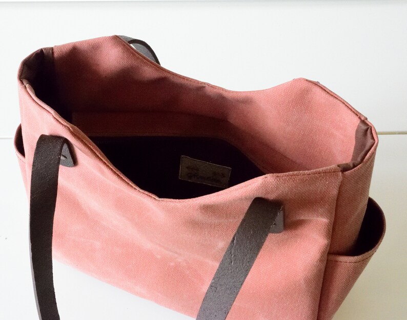K&ouml;nnte beinhalten: Eine rosafarbene Tragetasche mit dunkelbraunen Ledergriffen und -besatz. Die offene Tasche zeigt ein dunkles Innenfutter und Seitentaschen. Die Tasche hat ein schlichtes, klassisches Design.