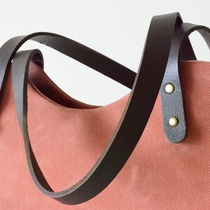 K&ouml;nnte beinhalten: Korallfarbene Tragetasche mit dunkelbraunen Ledergriffen. Die Griffe sind mit messingfarbenen Nieten am Beutel befestigt. Die Tasche hat ein schlichtes, klassisches Design, das f&uuml;r den t&auml;glichen Gebrauch geeignet ist.